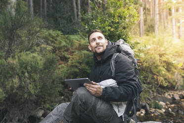 Lächelnder Mann sitzt mit digitalem Tablet im Wald - JCCMF04328