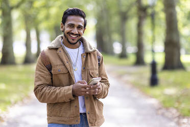 Smiling young man holding mobile phone in public park - WPEF05364