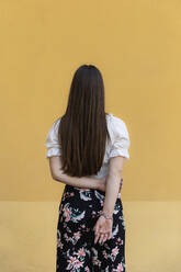 Teenage girl with hands behind back standing against yellow background - MRAF00645