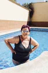 Smiling senior woman with hand on hip standing in swimming pool during vacation - EGHF00189