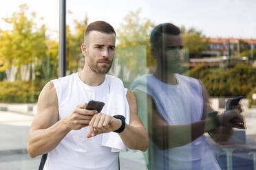 Sportler mit Mobiltelefon und Armbanduhr an Glaswand gelehnt - IFRF01141