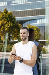 Smiling male athlete holding mobile phone in front of glass wall - IFRF01143