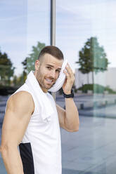 Smiling male athlete cleaning sweat with towel by glass wall - IFRF01146