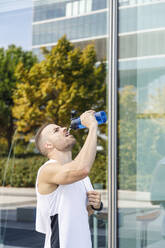 Male athlete drinking water by glass wall - IFRF01148