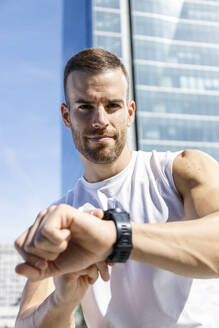 Lächelnder junger Sportler, der an einem sonnigen Tag seine Armbanduhr überprüft - IFRF01152