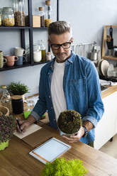 Man checking plant while writing on paper at kitchen counter - GIOF13883