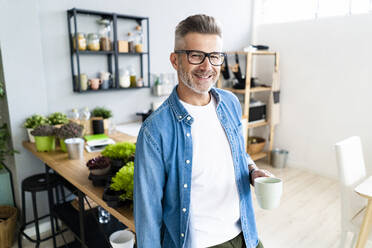 Smiling man wearing eyeglasses holding coffee cup at home - GIOF13894