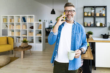 Man holding coffee cup while talking on mobile phone at home - GIOF13898