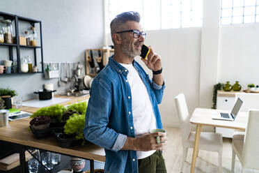 Älterer Mann hält Kaffeetasse, während er zu Hause mit seinem Smartphone telefoniert - GIOF13899