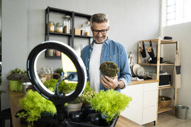 Smiling mature male influencer with potted plant vlogging at home - GIOF13902