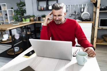 Mann mit Kopf in der Hand, der auf einen Laptop schaut, während er am Tisch sitzt - GIOF13907