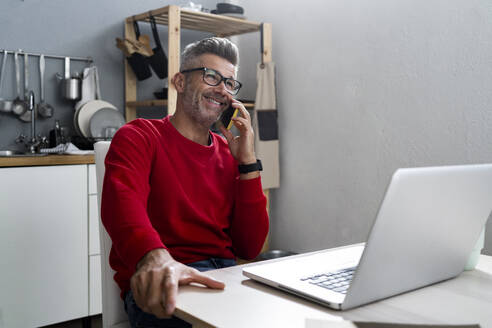 Lächelnder Mann mit Laptop, der zu Hause mit seinem Smartphone telefoniert - GIOF13913