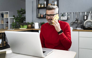 Älterer Mann mit Brille, der mit seinem Laptop am Tisch sitzt und mit seinem Smartphone spricht - GIOF13914