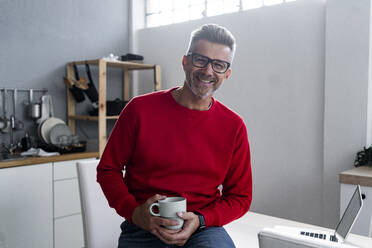 Smiling man with coffee cup siting at home - GIOF13916