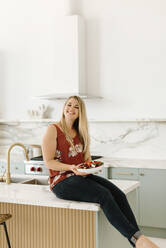 Smiling female nutritionist with plate sitting on kitchen counter - SMSF00577