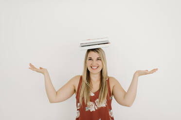 Glückliche Frau balanciert Bücher auf dem Kopf vor einer Wand - SMSF00596