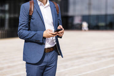 Businessman with backpack using mobile phone on footpath - WPEF05381