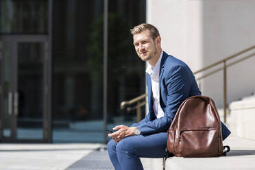 Lächelnder Geschäftsmann mit Mobiltelefon am Rucksack sitzend - WPEF05399