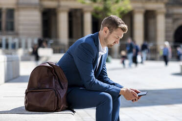 Male professional using mobile phone while sitting by backpack - WPEF05406