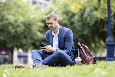 Männlicher Berufstätiger, der im Park sitzend eine Textnachricht über sein Mobiltelefon verschickt - WPEF05409