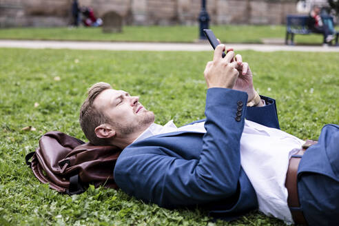 Männlicher Berufstätiger, der ein Mobiltelefon benutzt, während er im Park im Gras liegt - WPEF05414