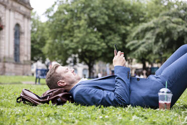 Junger Geschäftsmann, der ein Mobiltelefon benutzt, während er im Gras liegt - WPEF05415