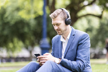 Businessman wearing wireless headphones using mobile phone in park - WPEF05422