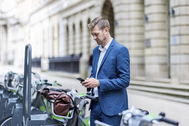 Junger Geschäftsmann, der an einer Fahrradabstellanlage sein Mobiltelefon benutzt - WPEF05432