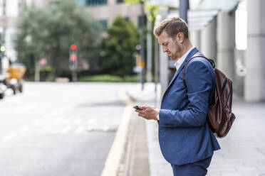 Young businessman with backpack using smart phone in city - WPEF05436