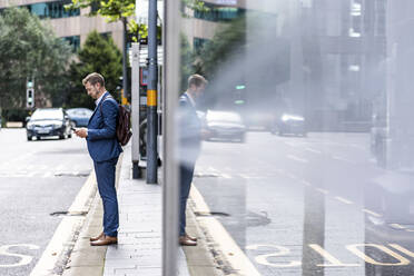 Businessman with backpack using mobile phone on footpath - WPEF05437