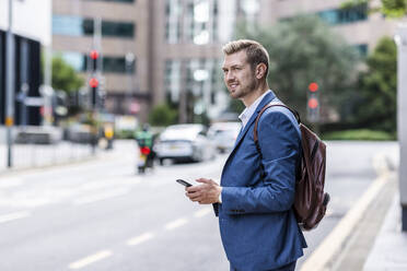 Smiling businessman wearing backpack holding mobile phone at city street - WPEF05439