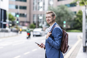 Young businessman with backpack holding smart phone in city - WPEF05440