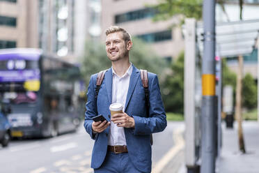 Smiling businessman holding disposable cup and mobile phone in city - WPEF05446