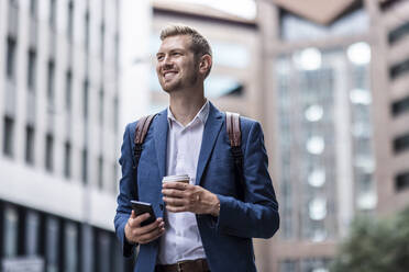 Happy young male professional with mobile phone and disposable cup in city - WPEF05447