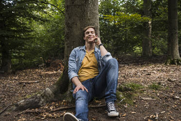 Smiling man with eyes closed leaning on tree trunk in forest - UUF24884