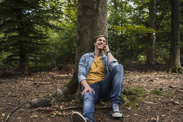 Happy man with hand on chin sitting by tree trunk in forest - UUF24885