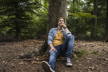 Thoughtful man with hand on chin sitting in forest - UUF24886