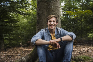 Happy man with brown hair sitting in front of tree trunk in forest - UUF24887