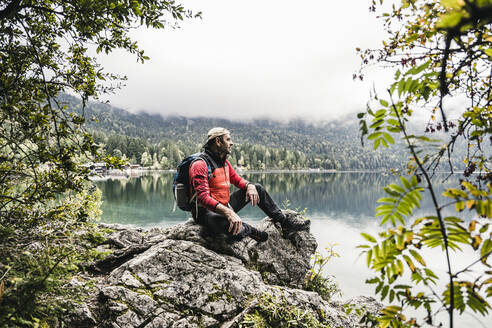 Männlicher Wanderer mit Rucksack auf einem Felsen im Wald sitzend - UUF24922