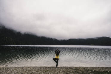 Woman practicing tree pose at lakeshore - UUF24936