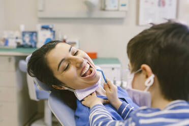 Neugieriger Junge in medizinischer Maske, der die Rolle des Zahnarztes spielt und die Zähne mit einem Zahnspiegel im Krankenhaus überprüft - ADSF31216