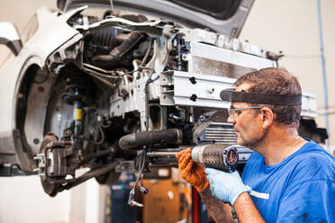 Seitenansicht eines fokussierten männlichen Mechanikers mit Werkzeug, der Metallteile eines kaputten Autos in der Reparatur schraubt - ADSF31217