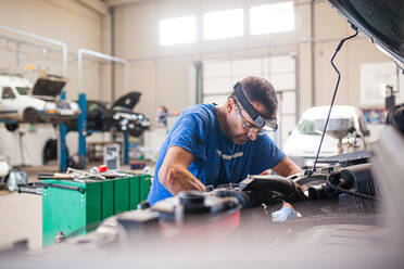 Seitenansicht eines beschäftigten reifen männlichen Technikers, der den Motor eines kaputten Autos in einer geräumigen Werkstatt repariert - ADSF31218