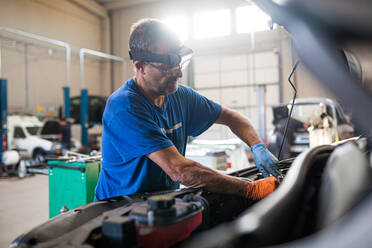 Seitenansicht eines beschäftigten reifen männlichen Technikers, der den Motor eines kaputten Autos in einer geräumigen Werkstatt repariert - ADSF31219