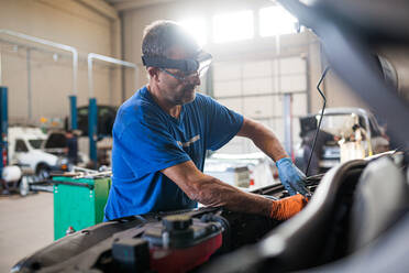 Side view of busy mature male technician fixing engine of broken car in spacious facility of repair service - ADSF31219