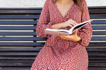 Cropped unrecognizable female in stylish clothes sitting with opened book on wooden bench against building with light wall in daytime - ADSF31225