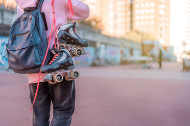 Crop unrecognizable female teenager wearing light pink hoodie and backpack with headphones and roller skates with oink shoelaces - ADSF31235