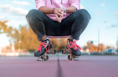 Ausgeschnittene, nicht erkennbare Frau, die einen hellrosa Kapuzenpulli, schwarze Jeans und Rollschuhe mit neonpinken Schnürsenkeln trägt und sich in einem Skatepark hinhockt - ADSF31238