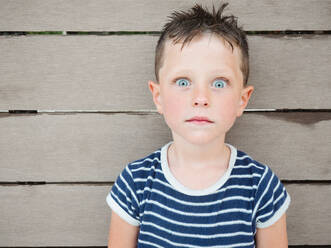 Amazed boy with wet hair wearing striped t shirt looking at camera with shocked expression against wooden wall - ADSF31264