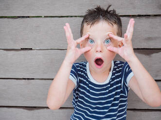 Playful amazed boy with wet hair opening eyes with hands while expressing astonishment and looking at camera - ADSF31265